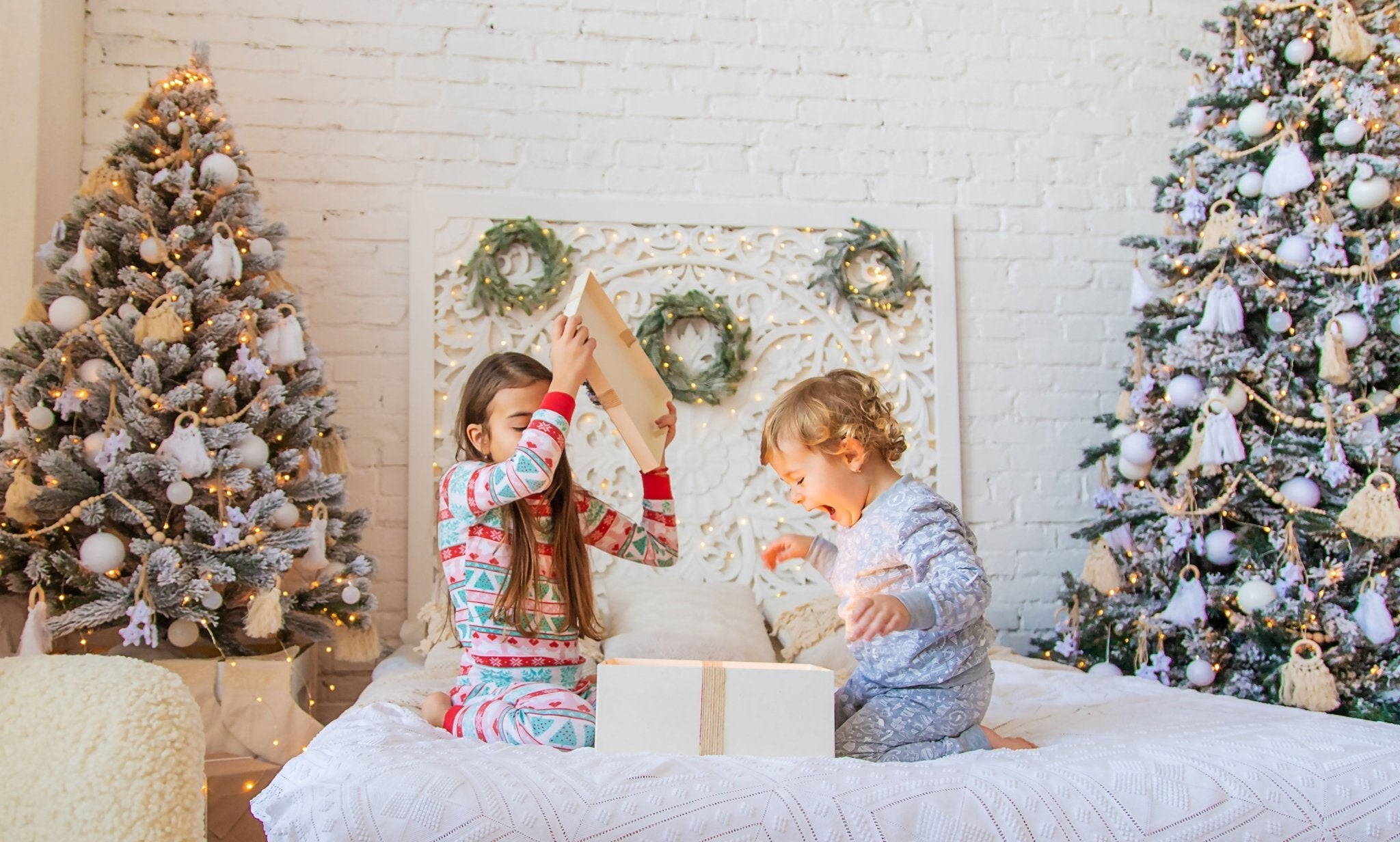 Zwei Kinder öffnen Weihnachtsgeschenke im Bett vor einem beleuchteten Weihnachtsbaum